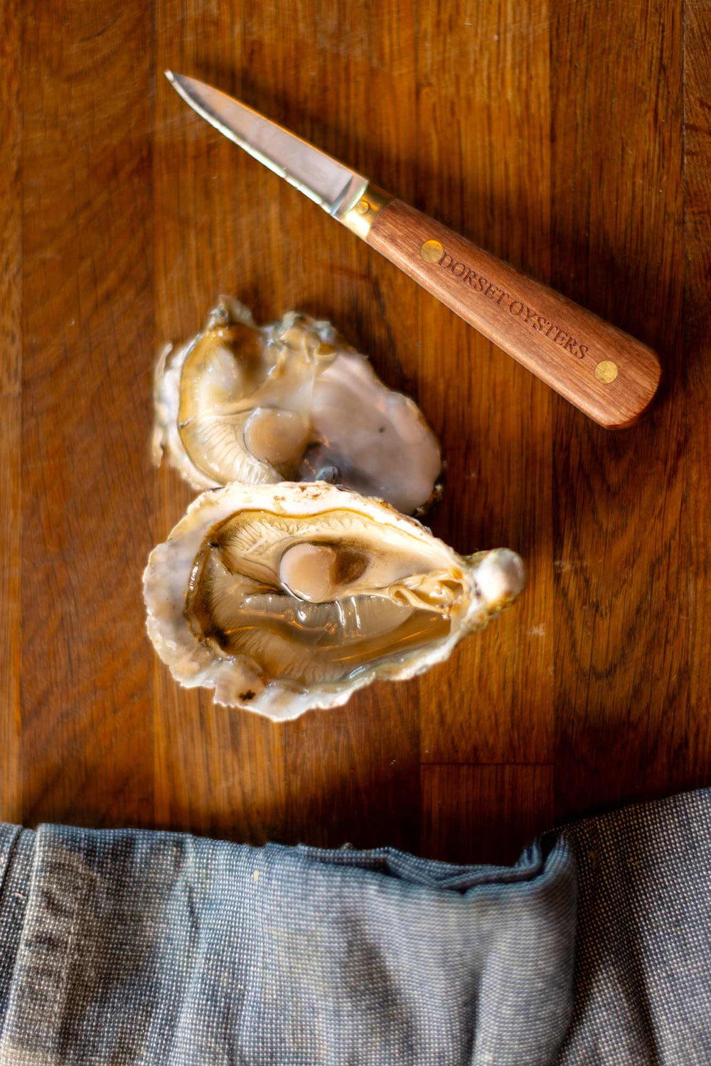 An open oyster next to a Dorset oysters oyster knife on a wooden board and a blue tea towel at the bottom of the image