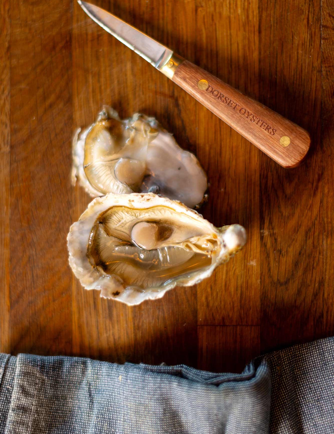 An open oyster next to a Dorset oysters oyster knife on a wooden board and a blue tea towel at the bottom of the image