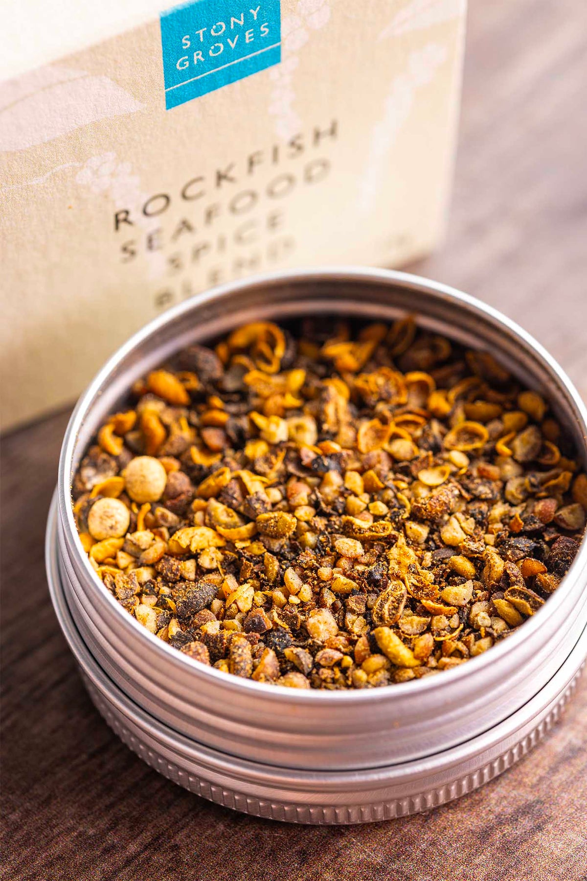 A round metal tin filled with a spice blend sits on a wooden surface, with the branded box blurred in the background. The visible text reads Stony Groves Rockfish Seafood Spice Blend.