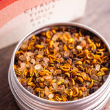 A close-up of a round silver tin filled with citrusy tincture mix from Stony Groves, with the branded box blurred in the background on a wooden surface.