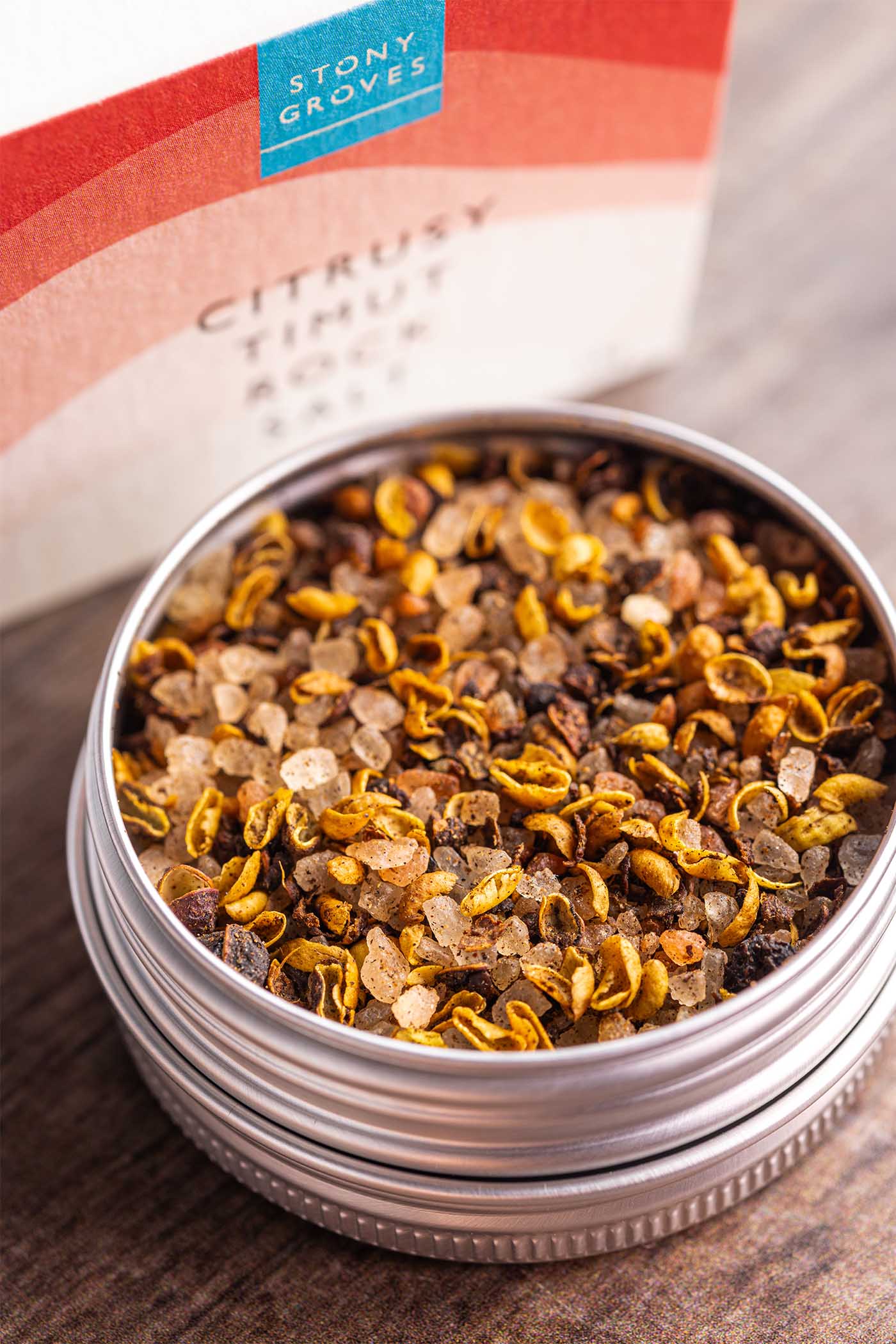 A close-up of a round silver tin filled with citrusy tincture mix from Stony Groves, with the branded box blurred in the background on a wooden surface.