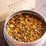 A round metal tin filled with a spice blend sits on a wooden surface, with the branded box blurred in the background. The visible text reads Stony Groves Rockfish Seafood Spice Blend.