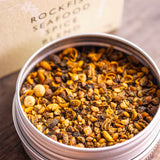 A round metal tin filled with a spice blend sits on a wooden surface, with the branded box blurred in the background. The visible text reads Stony Groves Rockfish Seafood Spice Blend.