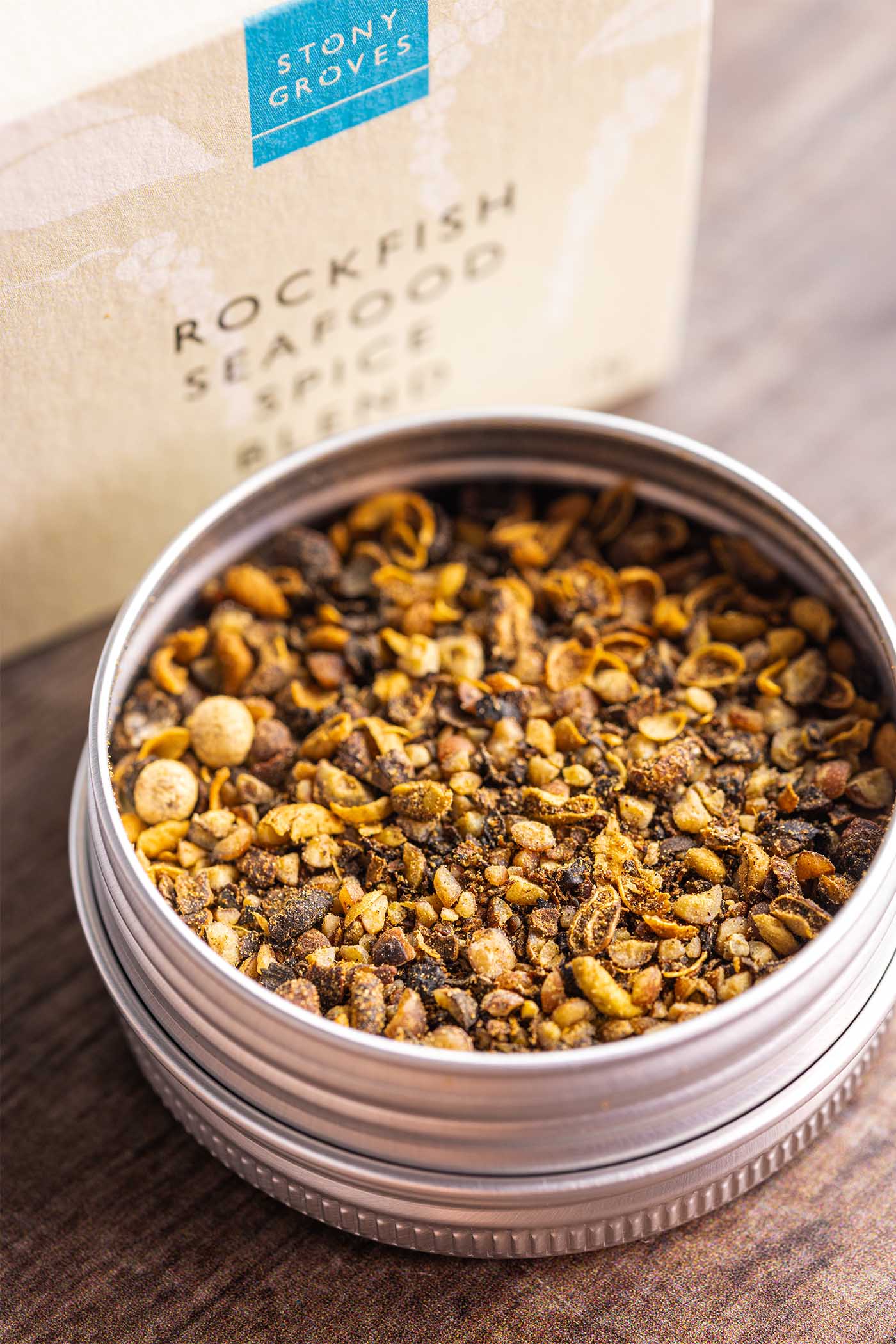A round metal tin filled with a spice blend sits on a wooden surface, with the branded box blurred in the background. The visible text reads Stony Groves Rockfish Seafood Spice Blend.