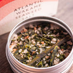 A close-up of a round metal tin filled with Stony Groves Atlantic Wakame Seaweed Salt, with the branded box blurred in the background on a wooden surface.