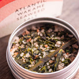 A close-up of a round metal tin filled with Stony Groves Atlantic Wakame Seaweed Salt, with the branded box blurred in the background on a wooden surface.