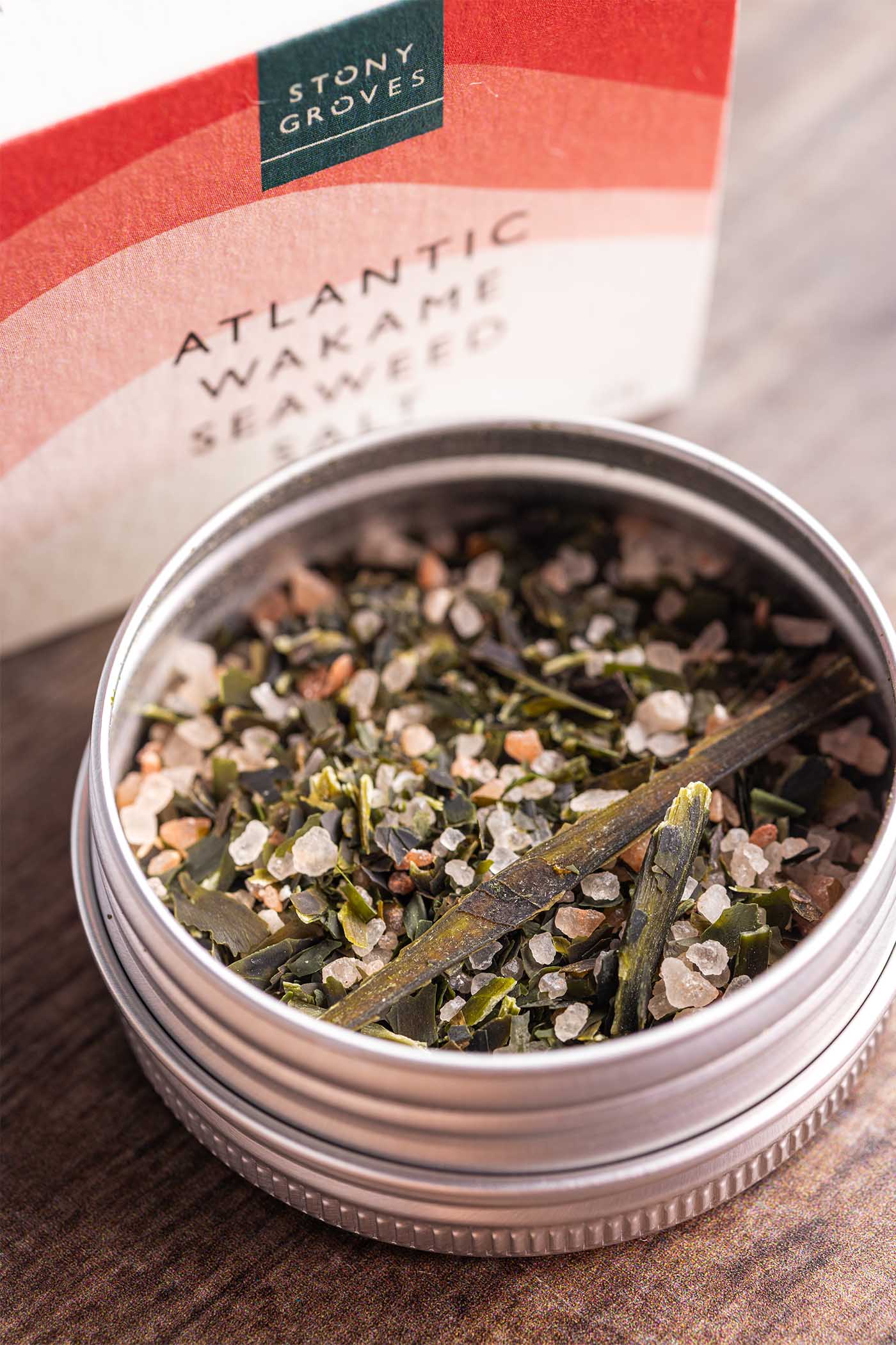 A close-up of a round metal tin filled with Stony Groves Atlantic Wakame Seaweed Salt, with the branded box blurred in the background on a wooden surface.