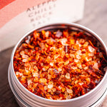 Stony Groves Aleppo Chilli Rock Salt in a round silver metal tin, shown open with coarse red and orange chili-infused salt crystals inside, with the branded box blurred in the background on a wooden surface.