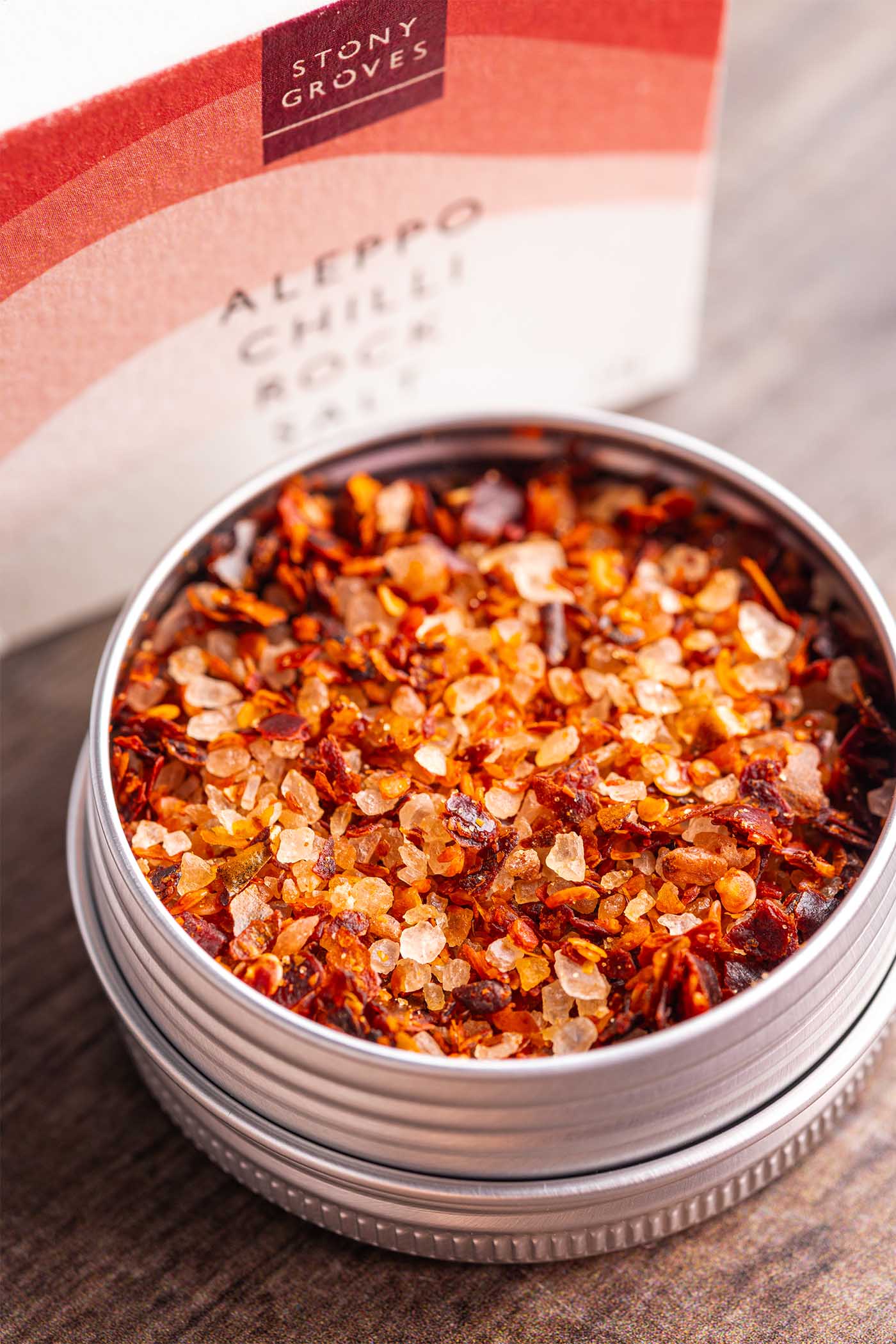 Stony Groves Aleppo Chilli Rock Salt in a round silver metal tin, shown open with coarse red and orange chili-infused salt crystals inside, with the branded box blurred in the background on a wooden surface.