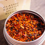 An open round metal tin filled with colorful loose-leaf spice blend, with the branded Stony Groves Classic Kampot Spice Blend box blurred in the background on a wooden surface.