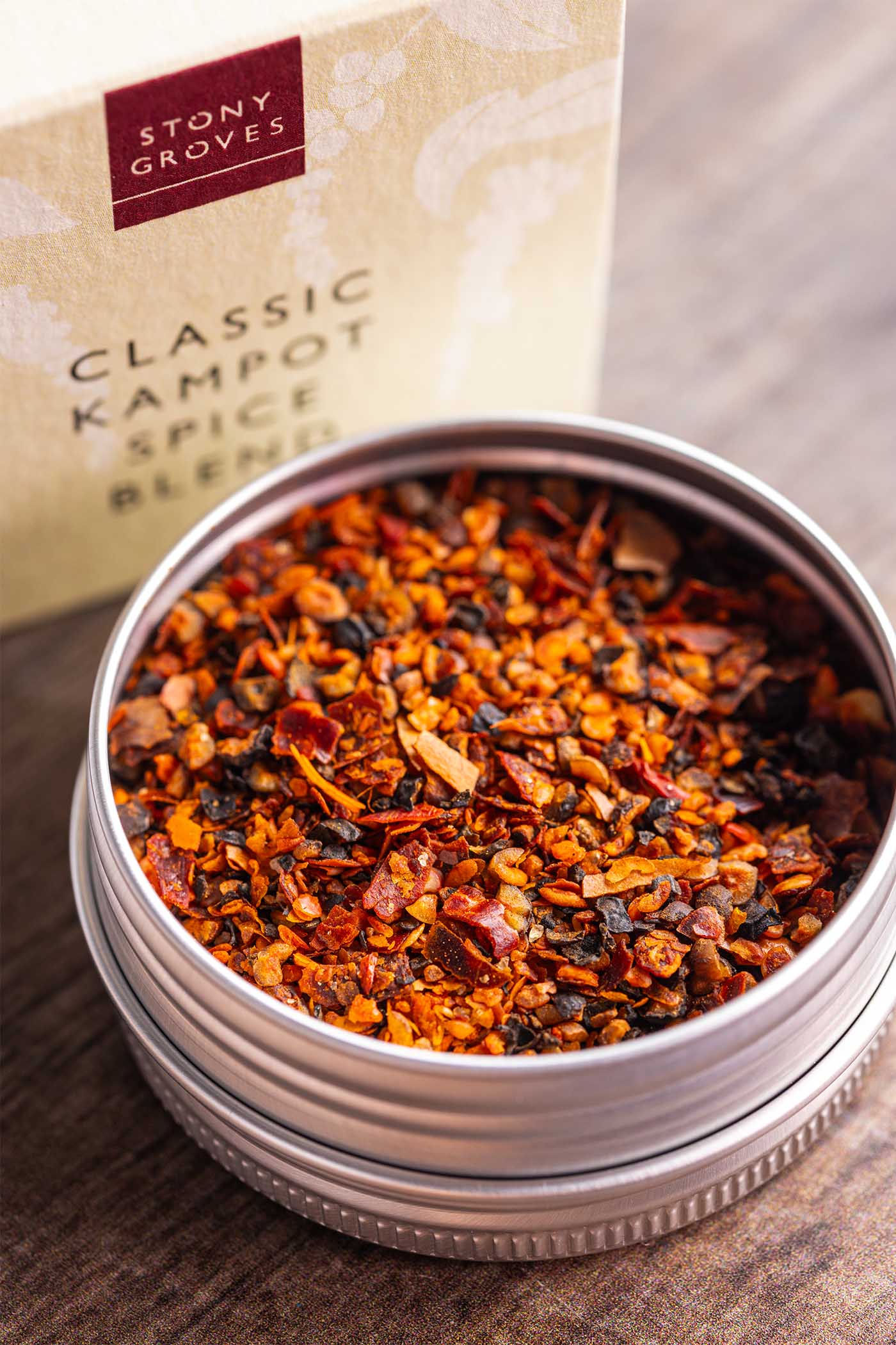 An open round metal tin filled with colorful loose-leaf spice blend, with the branded Stony Groves Classic Kampot Spice Blend box blurred in the background on a wooden surface.