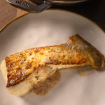 A close-up of a cooked fish fillet of Halibut resting on a white plate with a gold rim, with a dark cast iron skillet partially visible at the top. The skillet handle shows the embossed symbol 'No10' from Hornbeam cookware.