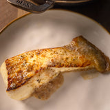 A close-up of a cooked fish fillet of Halibut resting on a white plate with a gold rim, with a dark cast iron skillet partially visible at the top. The skillet handle shows the embossed symbol 'No10' from Hornbeam cookware.