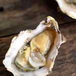 An open oyster shell filled with raw oyster meat on a rustic dark wooden surface, with another shell partially visible in the upper right.