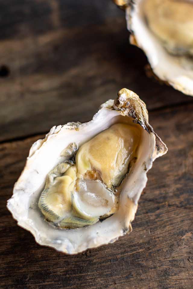 An open oyster shell filled with raw oyster meat on a rustic dark wooden surface, with another shell partially visible in the upper right.