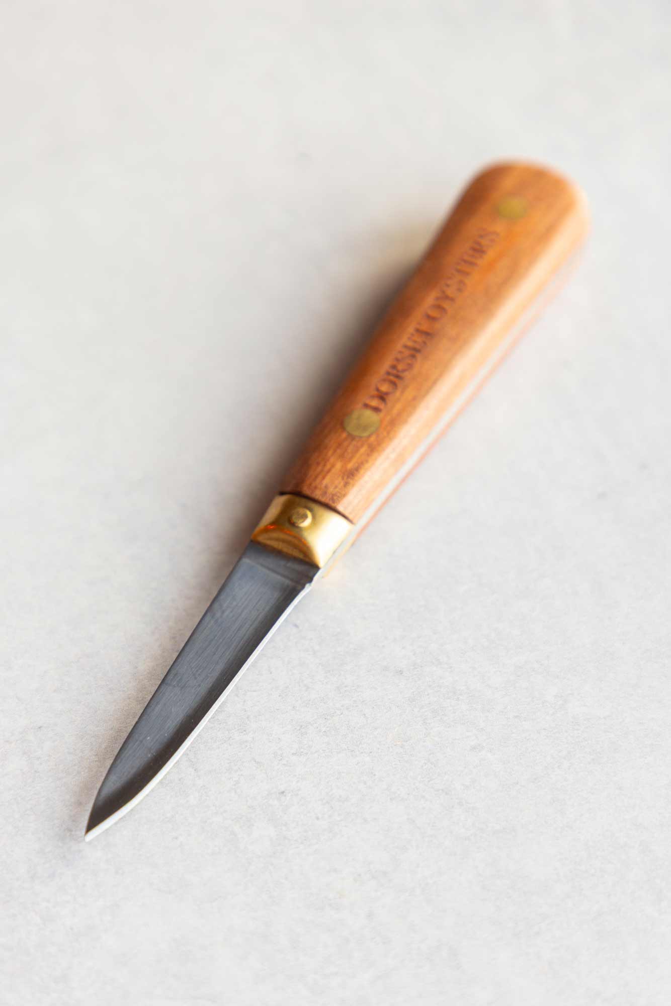 A small oyster knife with a light brown wooden handle and a dark steel blade, shown on a light gray textured background. The handle features the brand name 'Dorset Oysters' engraved vertically.