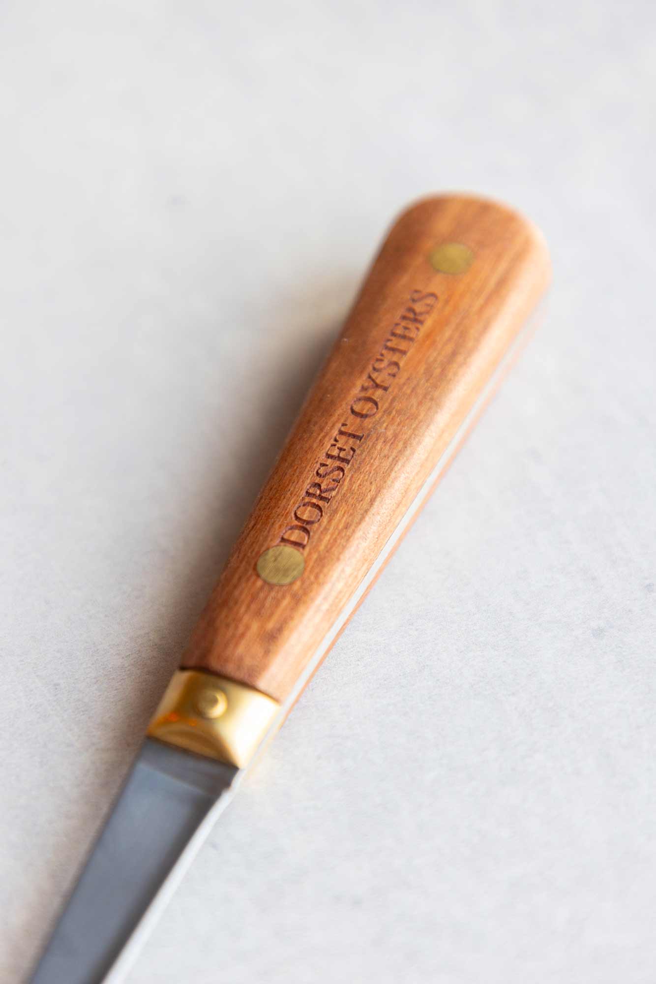 A close-up of a Dorset Oysters knife with a polished wooden handle engraved with the brand name, set against a light gray background.