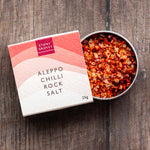 Aleppo Chilli rock salt open pot sitting half in its cardboard packaging on a wooden background. 