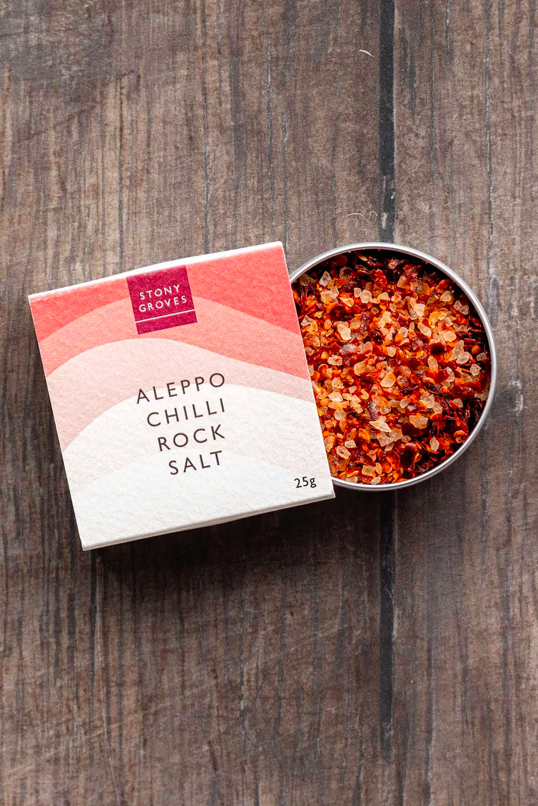 Aleppo Chilli rock salt open pot sitting half in its cardboard packaging on a wooden background. 