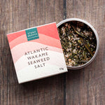 Atlantic Wakame Seaweed Salt by Stony Gloves, shown with its coral and cream gradient box next to an open round tin displaying the dried seaweed inside, set on a dark wood background.