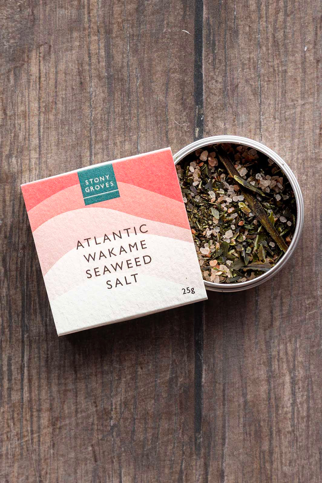 Atlantic Wakame Seaweed Salt by Stony Gloves, shown with its coral and cream gradient box next to an open round tin displaying the dried seaweed inside, set on a dark wood background.
