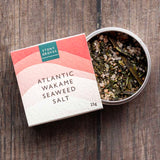 Atlantic Wakame Seaweed Salt by Stony Gloves, shown with its coral and cream gradient box next to an open round tin displaying the dried seaweed inside, set on a dark wood background.