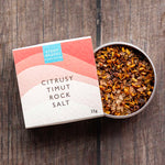 A tin of citrusy timut rock salt next to its paper packaging box on a dark wooden surface. The box is white with pink and peach wave graphics and a blue 'Stony Groves' logo.