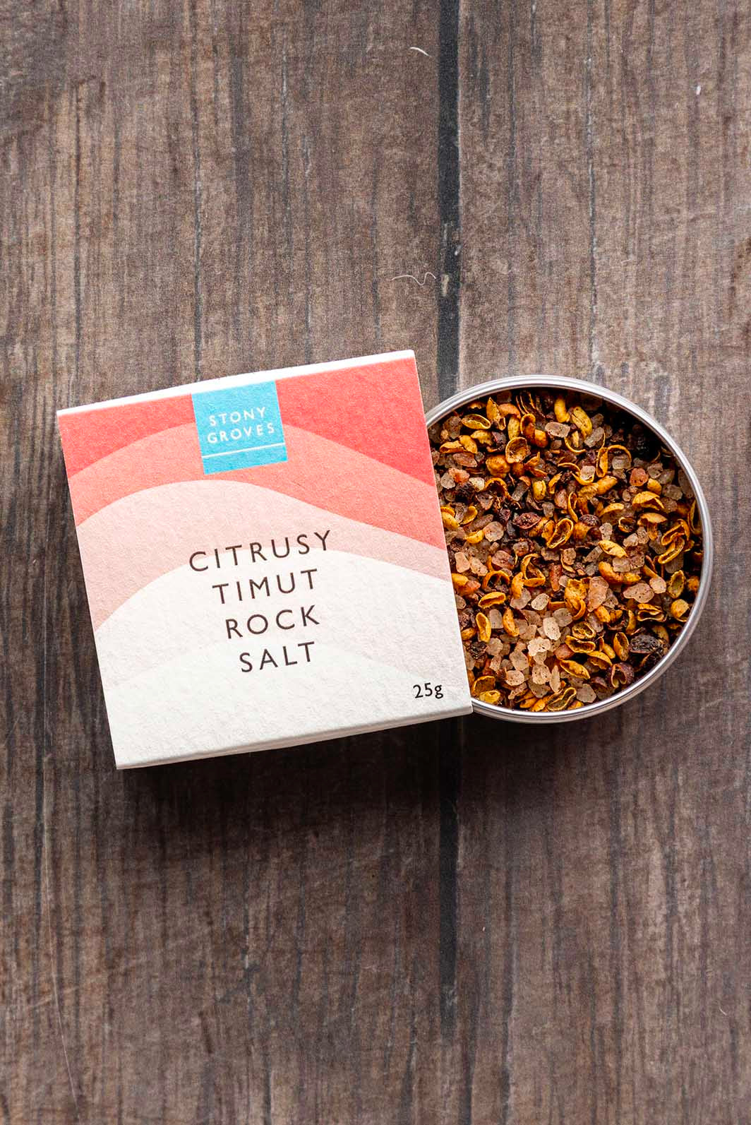 A tin of citrusy timut rock salt next to its paper packaging box on a dark wooden surface. The box is white with pink and peach wave graphics and a blue 'Stony Groves' logo.