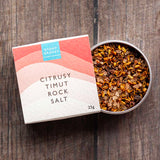 A tin of citrusy timut rock salt next to its paper packaging box on a dark wooden surface. The box is white with pink and peach wave graphics and a blue 'Stony Groves' logo.