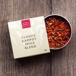 A 15g box of Stony Groves Classic Kampot Spice Blend next to an open metal tin filled with the colorful ground spice blend, set on a dark rustic wooden background.