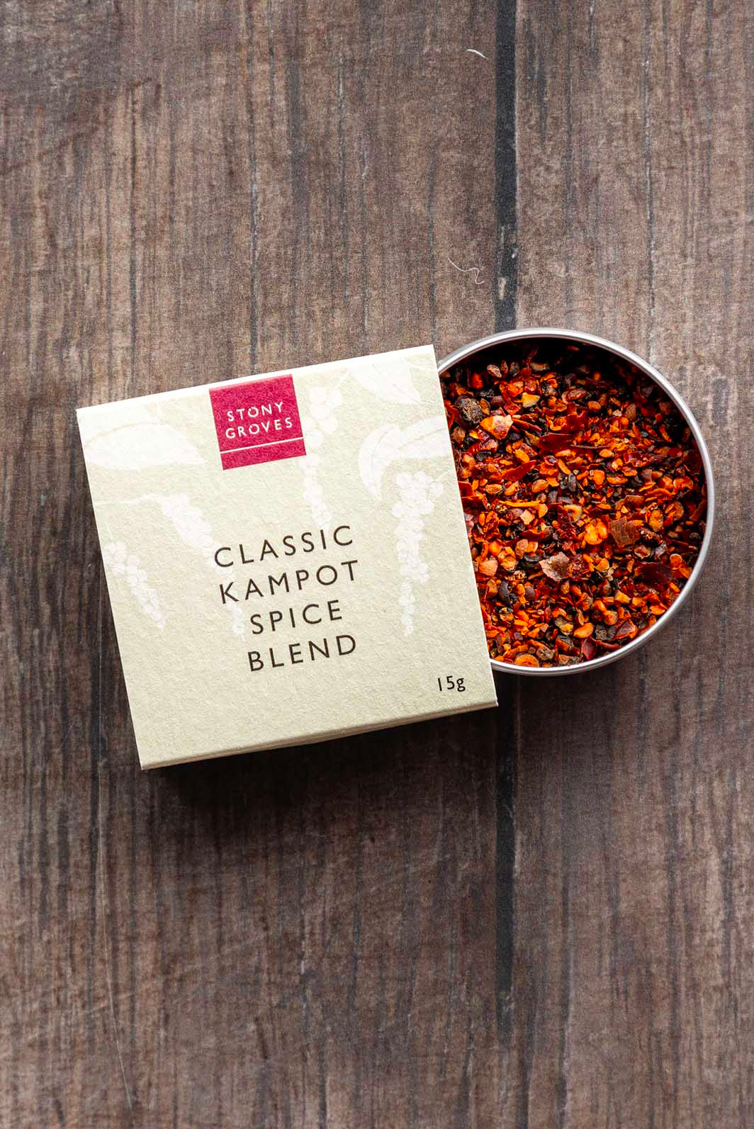 A 15g box of Stony Groves Classic Kampot Spice Blend next to an open metal tin filled with the colorful ground spice blend, set on a dark rustic wooden background.