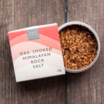 A 25g box of Stony Gloves Oak-Smoked Himalayan Rock Salt next to an open round tin showing the coarse salt crystals, set on a dark wood background.