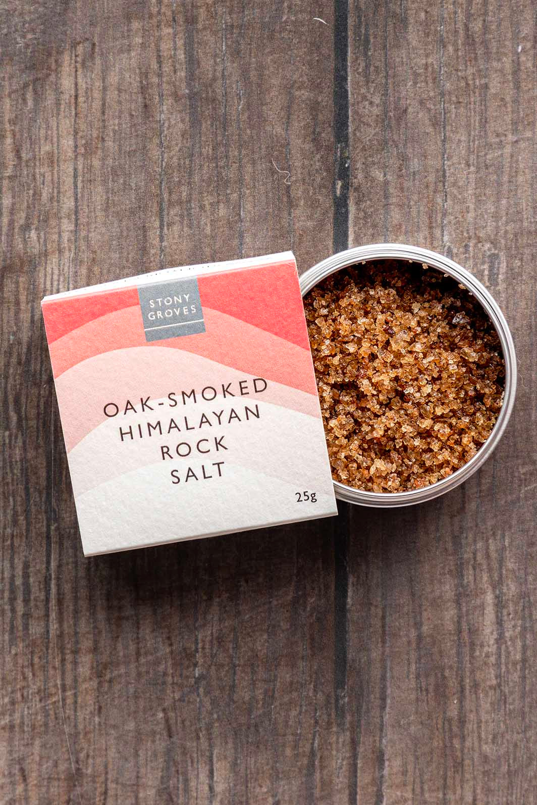 A 25g box of Stony Gloves Oak-Smoked Himalayan Rock Salt next to an open round tin showing the coarse salt crystals, set on a dark wood background.