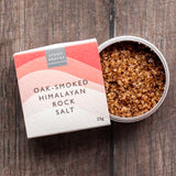 A 25g box of Stony Gloves Oak-Smoked Himalayan Rock Salt next to an open round tin showing the coarse salt crystals, set on a dark wood background.