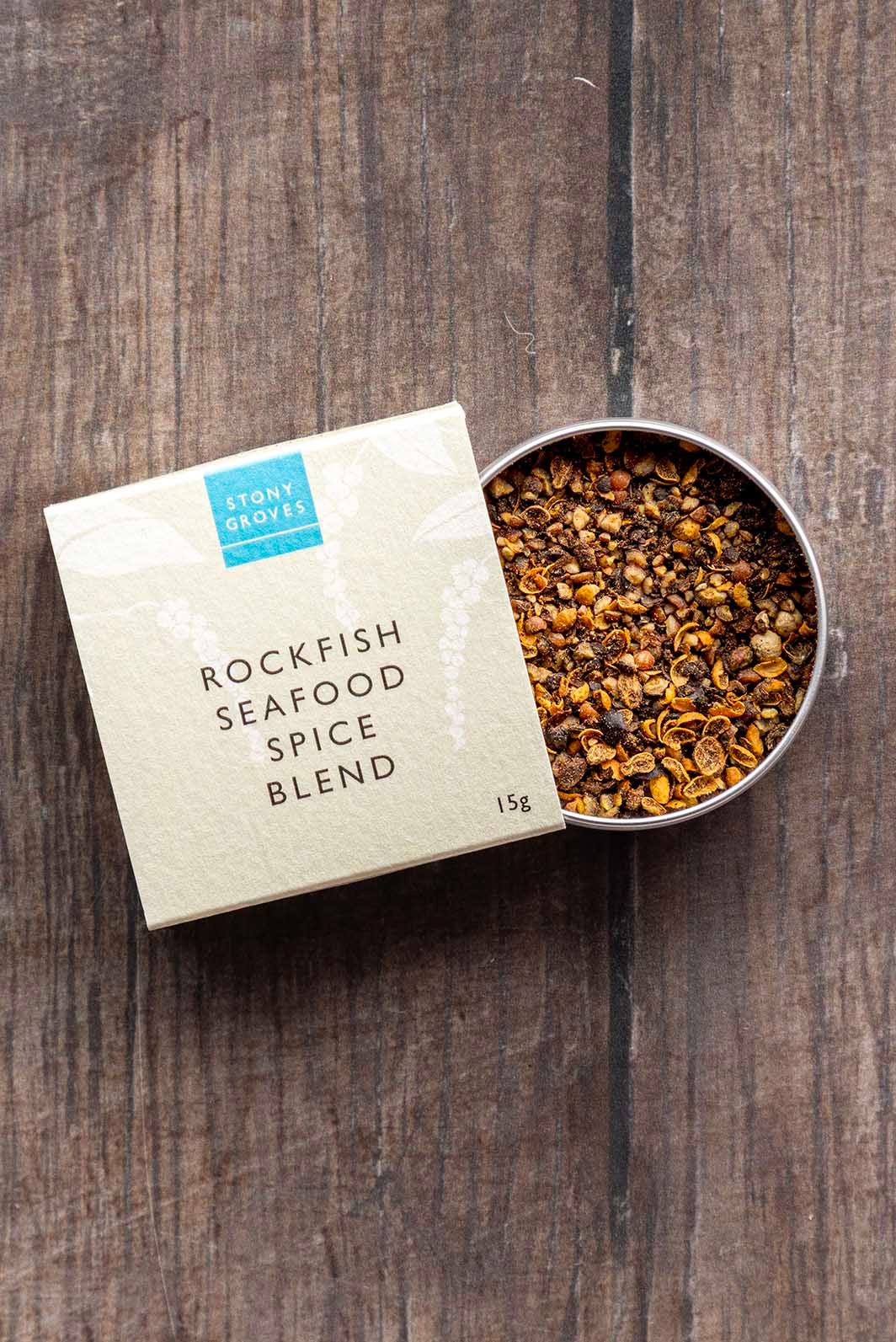 A square kraft paper box labeled 'STONY GROVES ROCKFISH SEAFOOD SPICE BLEND' sits beside an open round metal tin filled with ground spices, placed on a dark wood surface.