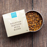 A square kraft paper box labeled 'STONY GROVES ROCKFISH SEAFOOD SPICE BLEND' sits beside an open round metal tin filled with ground spices, placed on a dark wood surface.