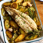 A whole Monkfish tail on a bed of roasted potatoes with rosemary in a roasting tray on a wooden table.