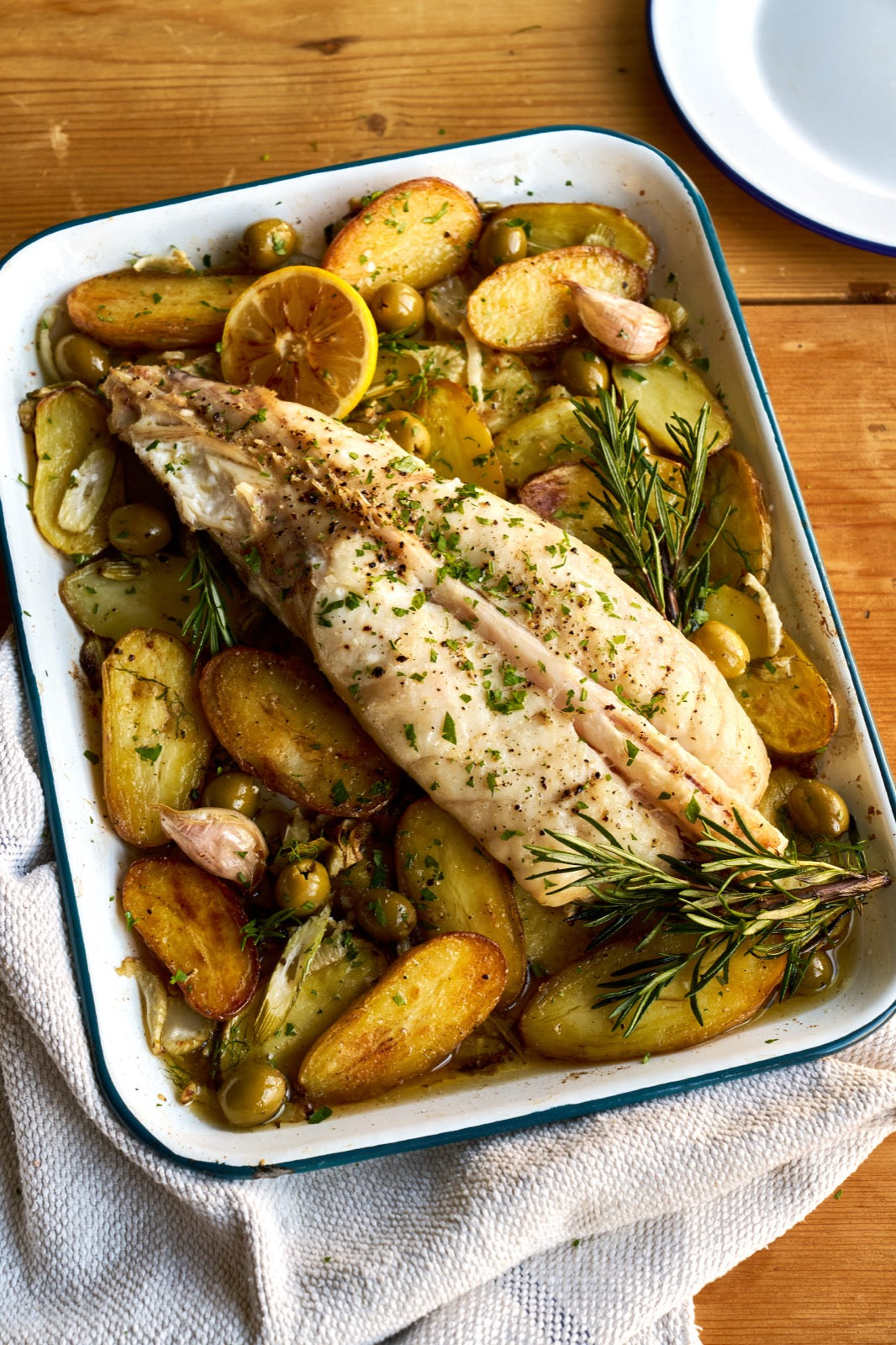 A whole Monkfish tail on a bed of roasted potatoes with rosemary in a roasting tray on a wooden table.