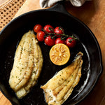 A portion of Brill fillets from Rockfish Online Seafood Market cooked in an iron skillet next to a wedge of lemon and some vine tomatoes