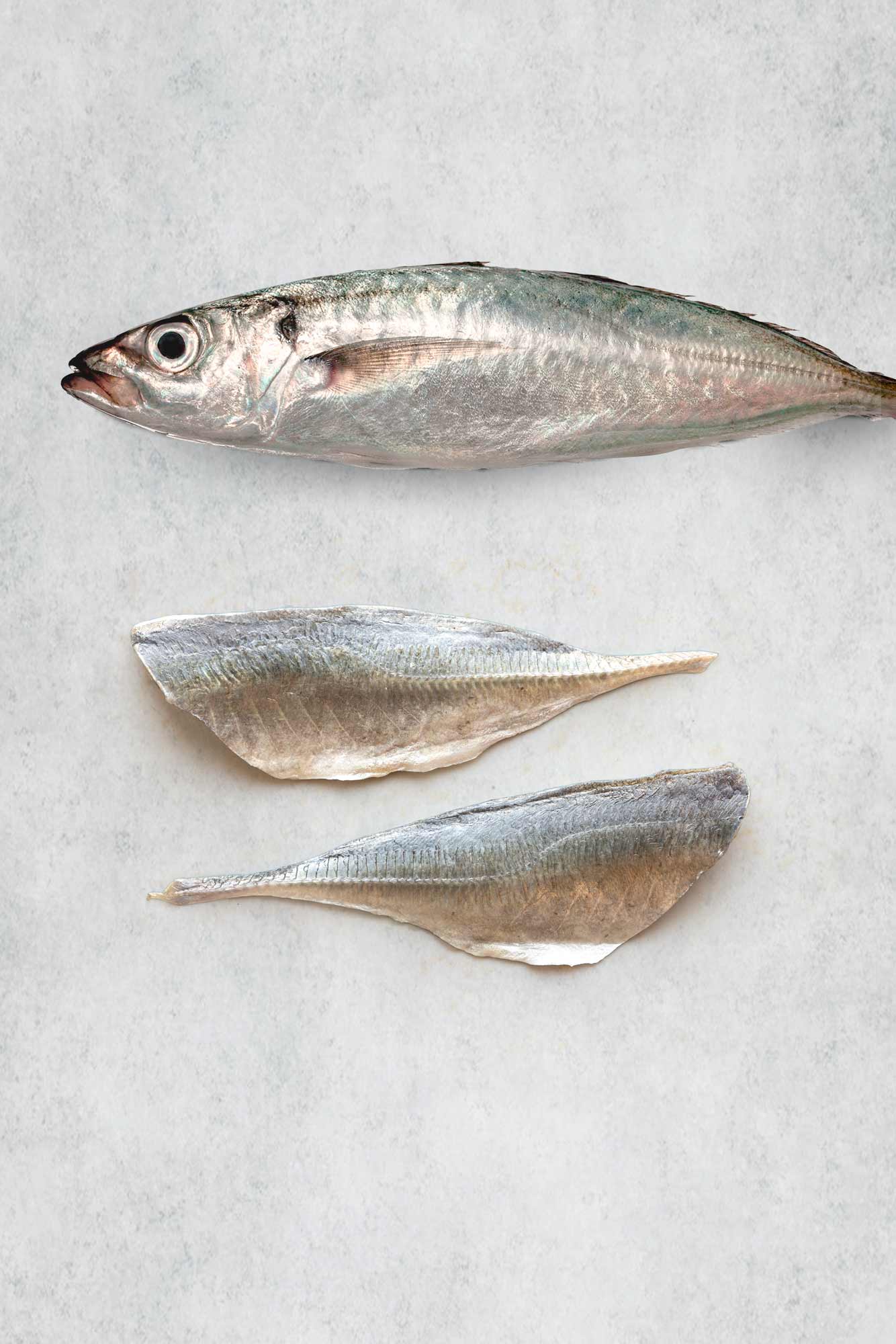 Two raw fish fillets of horse mackerel, including one whole horse mackerel sitting above it against a grey stone background.