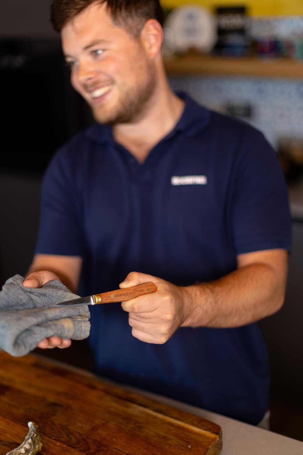 Tommy the fishmonger in a navy blue polo shirt holding a small oyster knife with a wooden handle and wiping it with a gray cloth in a kitchen setting.