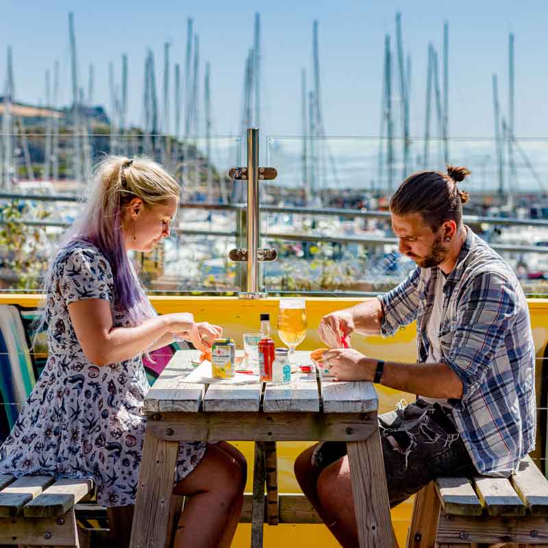 Two people enjoying Rockfish fish & chips with drinks in the sun at Plymouth Rockfish restaurant