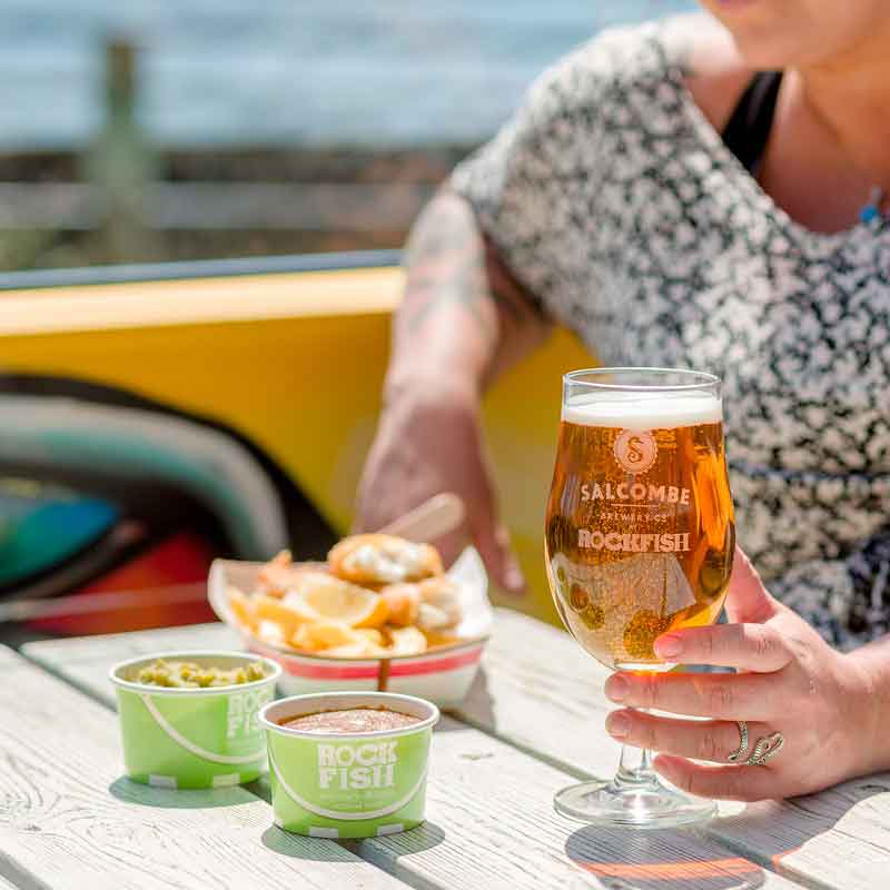 Woman enjoying portion of Rockfish takeaway fish and chips with sides & a drink outside in the sun at Rockfish Plymouth